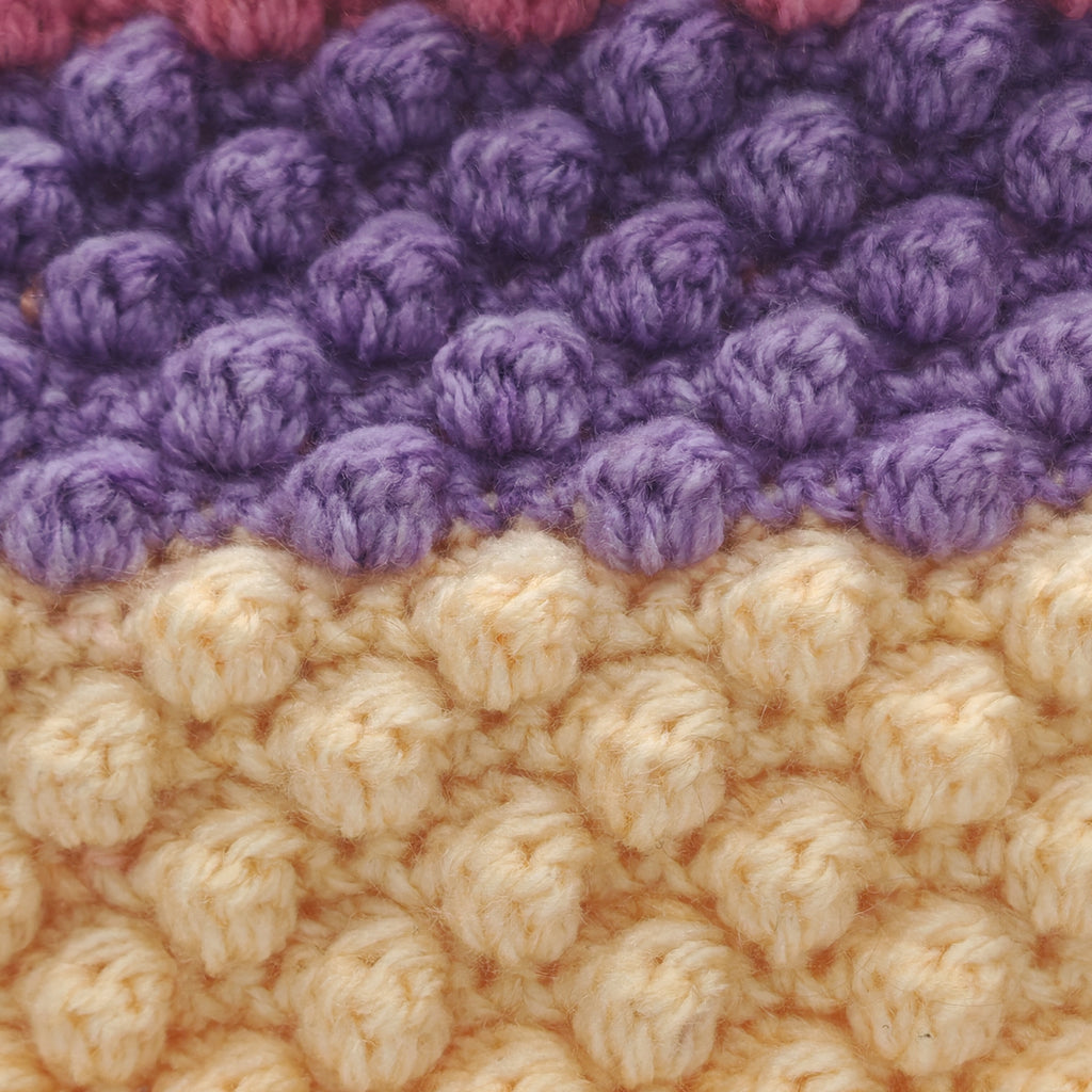 Detailansicht der Häkelstruktur eines Kissens mit Noppenmuster in Lila und Creme, weiche Garntextur