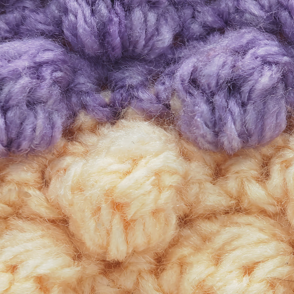 Makroaufnahme von gehäkelten Maschen in Lila und Creme, flauschige Garnstruktur und handgemachte Verarbeitung